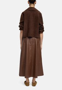 Osoba z upiętymi włosami nosi brązową zamszową kurtkę z podwiniętymi rękawami, brązową skórzaną spódnicę midi i jasnobrązowe buty wsuwane, stojąc tyłem.