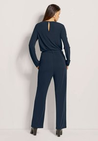 Navy jumpsuit met lange mouwen en een sleutelgatdetail aan de achterkant. Beschikt over een getailleerde taille en wijde broekspijpen, gemaakt van een gladde, rekbare stof.