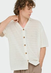 White knitted short-sleeve shirt featuring a V-neck, large brown buttons, and a loose fit with a textured pattern throughout.