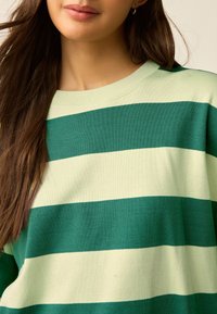 Bluză sport cu dungi verzi și crem, cu guler rotund și textură moale, având dungi orizontale largi.