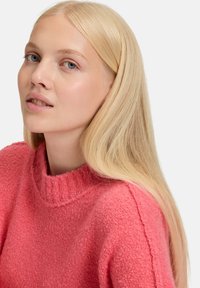 Helderroze brei-sweater met een hoge, geribbelde halslijn. Zachte, textuurstof met zichtbare stikseldetails. Glad, lang blond haar.