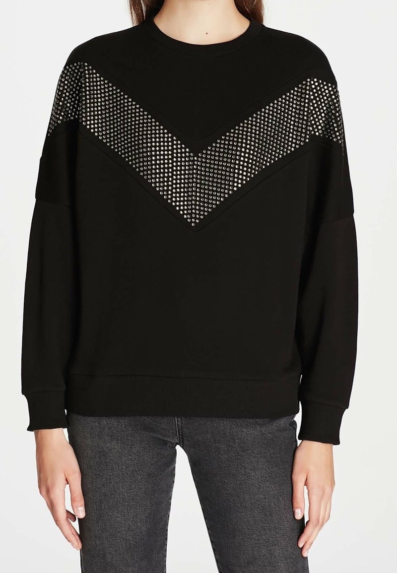 Femme portant un sweat-shirt noir à manches longues avec un détail chevron clouté argenté sur la poitrine et les épaules, assorti à un jean gris foncé.