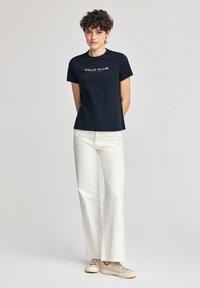 Marineblaues Baumwoll-T-Shirt mit "POLO CLUB"-Logo, kombiniert mit weißen weitgeschnittenen Jeans und beigen Sneakers. Einfaches, lässiges Design.