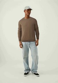 Sweater de malha castanho, jeans azul claro e sapatilhas pretas. A usar um chapéu cinza claro. Aposentado contra um fundo neutro.