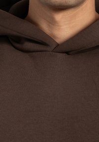 Närbild av en persons nacke som bär en mörkbrun huvtröja med en vikbar huva.