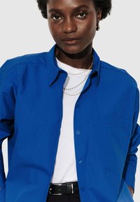 Femme portant une chemise bleu vif à boutons sur un haut blanc, avec des colliers superposés et une ceinture noire, contre un fond uni.