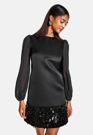 Mujer con vestido negro de mangas largas transparentes y falda con lentejuelas, mirando hacia abajo con el pelo lacio y oscuro y pendientes grandes y redondos.
