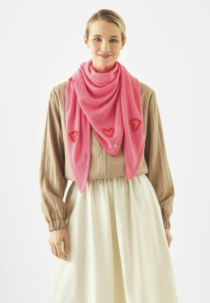 Frau mit blondem Haar trägt eine hellbraune Bluse, einen weißen Rock und einen rosa Schal mit roten Herzmustern, der um ihren Hals drapiert ist.