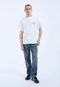 Bílý bavlněný tričko s grafickým potiskem na hrudi, spárované s modrými džíny a černými teniskami s bílými pruhy.