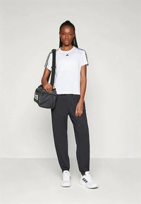 Λευκό T-shirt Adidas με μαύρο λογότυπο, μαύρο παντελόνι φόρμας και λευκά παπούτσια με ρίγες. Συνοδεύεται από μια μαύρη τσάντα γυμναστηρίου.