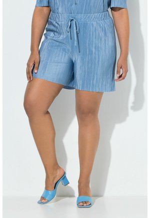 Vrouw die lichtblauwe, gestructureerde korte broek draagt met een trekkoord in de taille en bijpassende blauwe open teen blokhaksandalen, staand tegen een witte achtergrond.