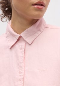 Lyserosa skjorte med krage laget av stoff med myk tekstur, med knappeåpning og brodert logo på venstre side.