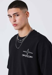 Camiseta negra oversize con el logo blanco "PXP" y diseño de estrellas. Lleva puestas cadenas de plata en capas. Corte de pelo corto y ordenado.