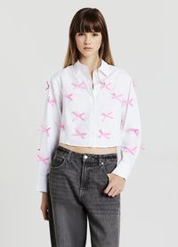 White cropped button-up shirt with pink bow accents, featuring a relaxed fit and long sleeves, worn with high-waisted dark gray jeans.