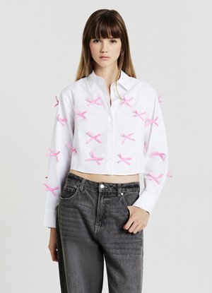 Camicia bianca cropped con dettagli a fiocco rosa, caratterizzata da una vestibilità rilassata e maniche lunghe, abbinata a jeans grigi scuri a vita alta.