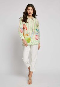 Multicolore blouse met knopen en een groen gestreept patroon, met illustraties van fruit en drankjes; gecombineerd met witte rechte pantalon.