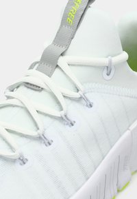 Chaussure de sport gris clair avec un tissu texturé, des lacets blancs et un accent vert vif sur la semelle. Elle présente une languette au dos pour un enfilage facile.