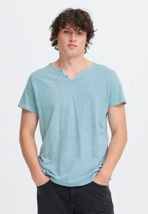 Jeune homme aux cheveux bouclés foncés, portant une chemise bleu clair à manches courtes et un pantalon noir, debout les mains dans les poches sur fond blanc.