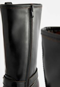 Schwarze Lederstiefel mit einem seitlichen Reißverschluss, akzentuiert mit braunen Nähten und einem Gurt um den Knöchel. Glatte Oberfläche und robuste Bauweise.