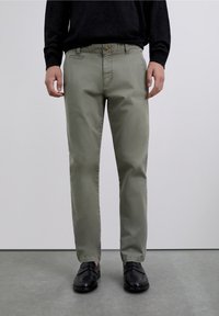 Pantalones chinos de color verde oliva claro hechos de algodón, con diseño de pierna recta, bolsillos delanteros y cierre de botón. Usados con zapatos negros.