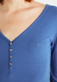 Blaues, geripptes Langarmoberteil mit V-Ausschnitt, ausgestattet mit fünf Holzknöpfen und einem gestickten "Lee"-Logo auf der linken Brust.