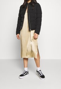 Giacca a vento nera con zip frontale, abbinata a un vestito slip dorato lucido con fessure laterali, e sneakers nere con suole bianche.