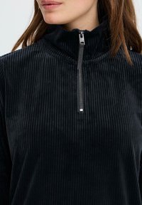 Zwarte corduroy pullover met hoge kraag, voorzien van een ritsluiting, geribbelde textuur en zachte stof. Afgemaakt met een zilveren ritslip.