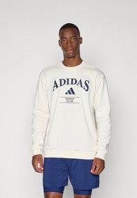 Krēms krāsas džemperis ar tumši zilu "ADIDAS TENNIS CLUB" druku, ribotam kakla izgriezumam un aprocēm, kopā ar tumši zilām biksēm. Mīksts audums, regulāra piegriezuma.