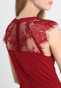 Femme portant une robe rouge sans manches avec des détails en dentelle florale et en mesh au niveau du haut du dos et des épaules.