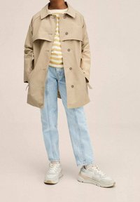 Beżowy trencz z dużym kołnierzem, szczegóły guzikowe, paskowana żółto-biała koszula, jasnoniebieskie dżinsy oraz beżowe sneakersy z akcentami.