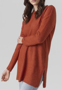 Femme portant un pull en maille à manches longues couleur rouille avec des fentes sur les côtés, associé à un jean bleu clair sur un fond uni.