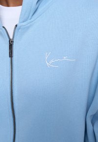 Light blue zip-up hoodie made of soft fabric. Features a silver zipper and white embroidered "Kany" logo on the chest. Casual design.