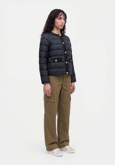Modèle portant une veste noire matelassée à boutons dorés, un pantalon cargo olive et des baskets blanches sur un fond de studio uni.