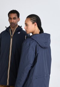 Vestes étanches de couleur marine avec capuches, présentant une fermeture éclair sur le devant et des rayures de couleur contrastante, exhibées sur deux modèles sous différents angles.