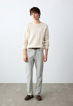 Mlad moški stoji ob beli steni, oblečen v krem pulover, svetlo sive kavbojke, rjav pas in rjave čevlje na sivi tleh.