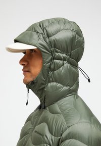 Green puffy jacket with a fitted hood, quilted texture, and a white brimmed cap. Features adjustable toggles at the collar.