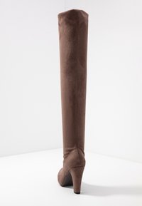 Bota marrón de ante por encima de la rodilla con un tacón afinado. Textura suave, diseño ajustado y una silueta de caña alta.