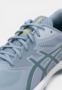 Lys blå atletisk sko med mesh-materiale, teksturerte mønstre, reflekterende detaljer og gummisåle med en fremtredende logo.