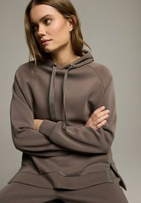 Brązowy hoodie o luźnym kroju, z kapturem ze sznurkiem, ściągaczami przy mankietach i bocznymi rozcięciami. Gładka tekstura i lekki materiał.