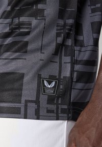 Nærbillede af sort og gråt mønstret stof med et patch, der står "Authentic Castore Product" på en persons side, der bærer hvide bukser.