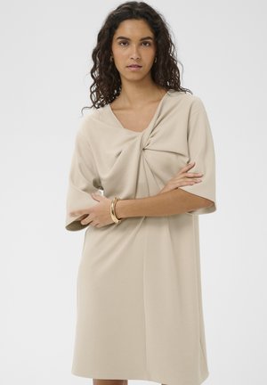 Käharate juustega naine, kes kannab beežikat kleiti, millel on keerutatud esiosa detail, kuldne käevõru ja rõngakujulised kõrvarõngad, seisab käed rüpes.