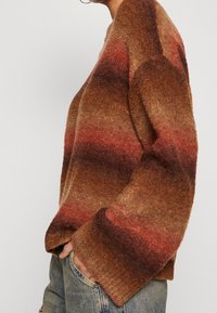 Suéter a rayas en tonos marrón y óxido con cuello redondo, puños acanalados y tejido texturizado, combinado con jeans de color azul claro.