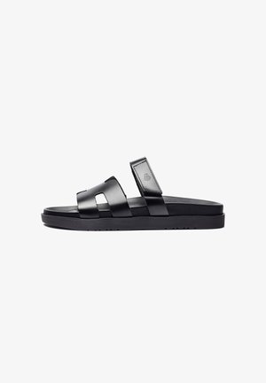 Sandale noire en cuir à enfiler avec deux larges brides, dont une avec une fermeture Velcro et un logo embossé, semelle plate.
