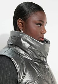 Metallisch silberne gepolsterte Weste mit hohem Kragen, ausgestattet mit einem gesteppten Design und strukturiertem Stoff. Getragen über einem schwarzen gemusterten Oberteil.