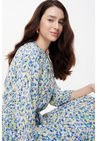 Abito floreale in blu, giallo e verde con maniche lunghe, scollo arricciato e cintura in vita. Tessuto leggero con una silhouette fluida.