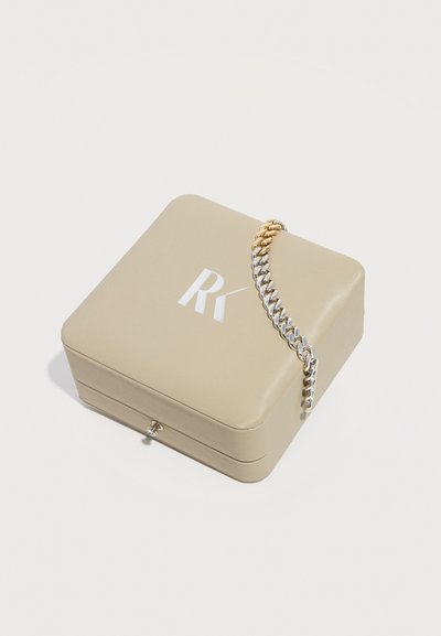 Μπεζ τετράγωνο κουτί κοσμημάτων με το λογότυπο "RK", που φέρει μια δίχρωμη αλυσίδα σε χρυσό και ασημί κρεμασμένη πάνω στη γωνία.