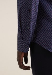 Camicia blu marina con piccoli motivi geometrici rosa, dotata di polsini abbottonati e una texture di tessuto liscia. Mano visibile di lato.