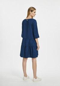 Granatowa sukienka do kolan z rękawami o długości trzech ćwiartek, z warstwonymi falbanami na końcu i guzikiem z tyłu. Miękka tekstura materiału.