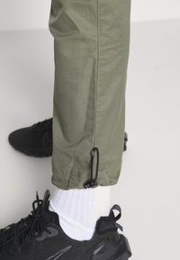 Kakifärgade cargobyxor med justerbara stålar, tillverkade av ett lätt material. I kombination med svarta sportskor och vita ribbade strumpor.