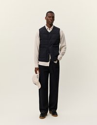 Άνδρας φορώντας ένα μαύρο ραφτό γιλέκο πάνω από κρέμ μακρυμάνικο πουκάμισο, μαύρες φαρδιές παντελόνες, κρατώντας ένα μπεζ καπέλο με το ένα χέρι.